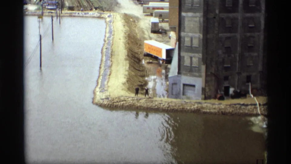 Final 1969 Saint Paul frame shows two distant people on a levee beside floodwater, near a large building, industrial structures, and a parked box truck.