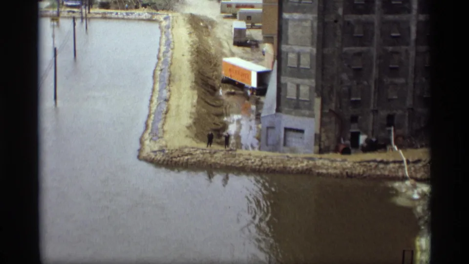 Saint Paul riverfront flooding in 1969, with two distant figures near a warehouse and a trailer close to the waterline.
