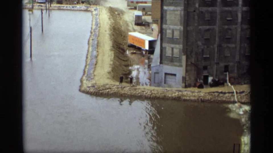 1969 home-movie view of Saint Paul floodwater along an industrial embankment with a warehouse, trailer, and two figures.