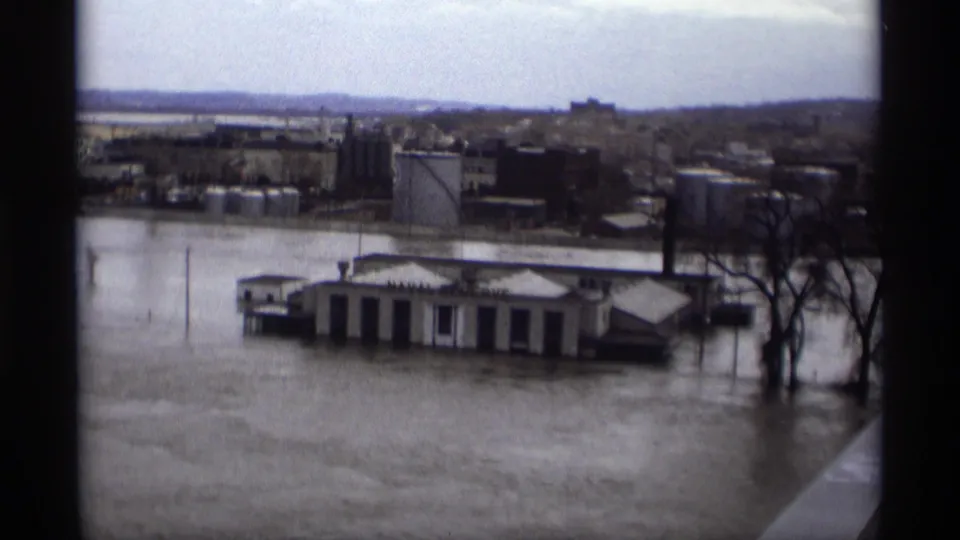 1969 Saint Paul flood scene with low waterfront buildings and bare trees partly surrounded by muddy water.