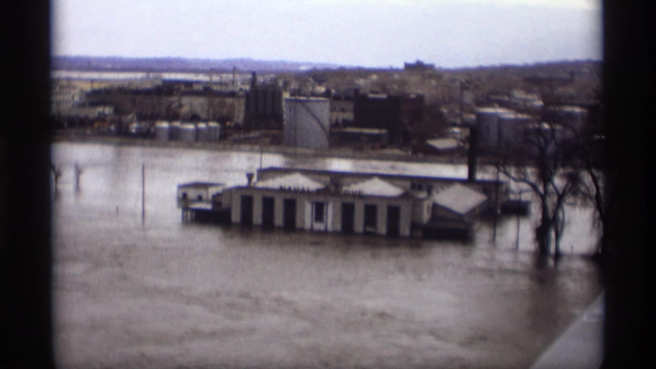 1969 Saint Paul floodwater covers a riverfront area around a low building, with industrial buildings and bare trees in the distance.