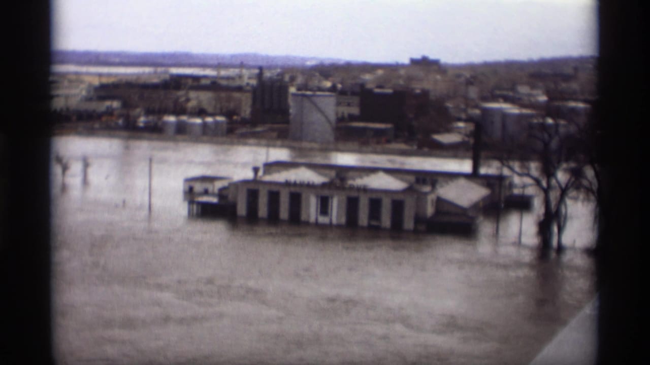 A partially submerged riverfront building sits in floodwater near bare trees and distant buildings.