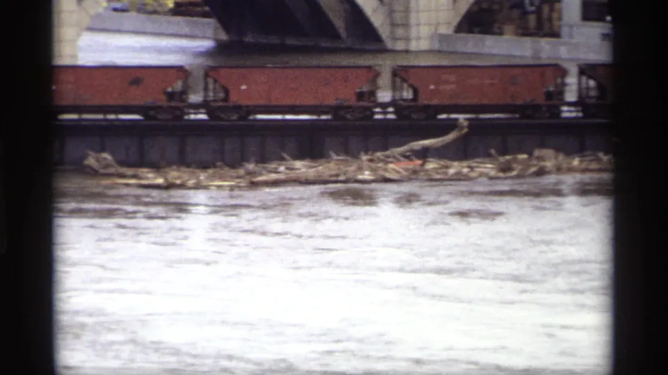 Red freight cars on a bridge over a slow river in the Saint Paul industrial area, with debris gathered along the shoreline.