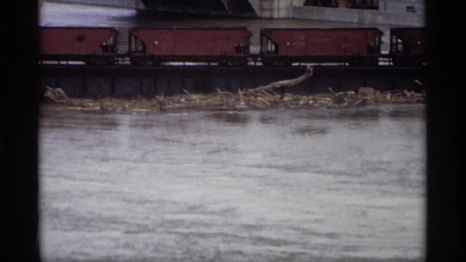 Saint Paul industrial river view with red freight cars crossing a bridge and driftwood or debris collected below.