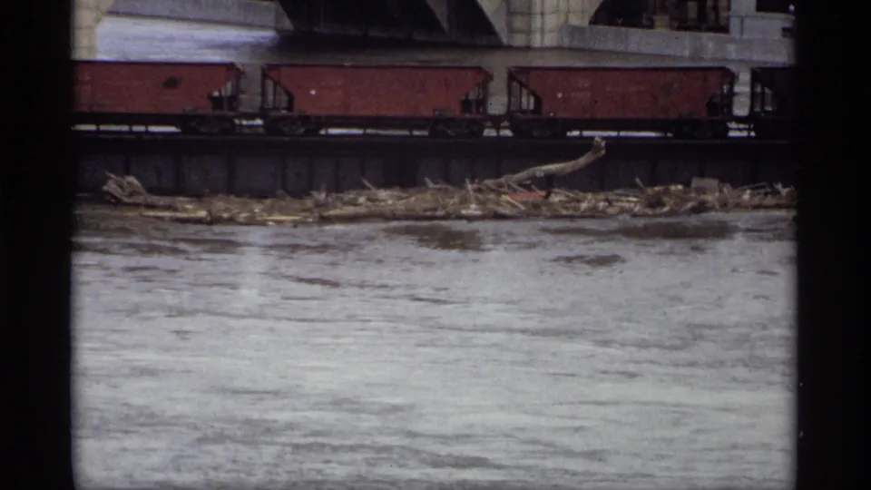 1969 Saint Paul riverfront frame showing red freight cars on a bridge above slow water and accumulated debris along the bank.