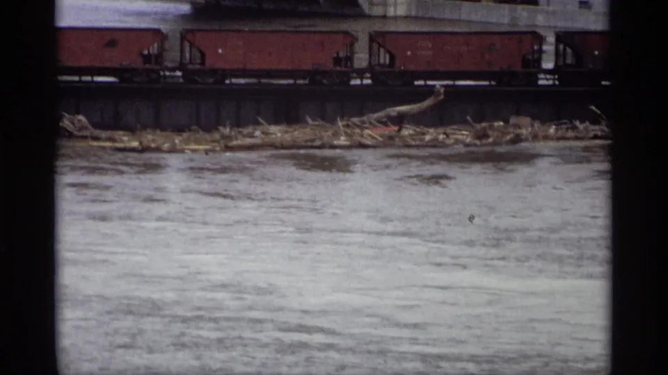 1969 Saint Paul 8mm frame showing red freight cars on an elevated track above murky river water and shoreline debris.