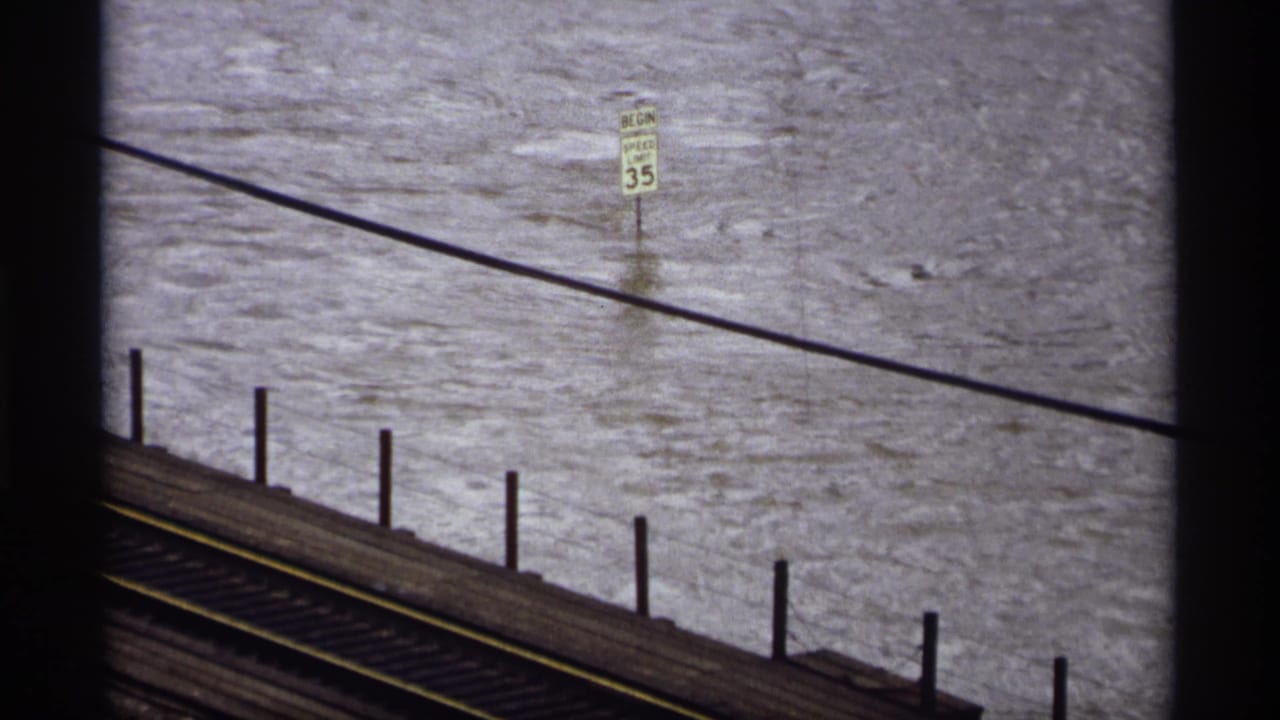 Saint Paul 1969 home-movie frame showing turbulent floodwater beside railroad tracks and a partially submerged speed limit sign.