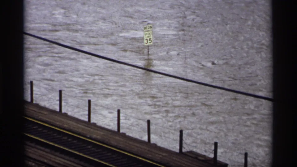Saint Paul 1969 home-movie frame showing turbulent floodwater beside railroad tracks and a partially submerged speed limit sign.