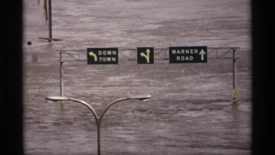 Saint Paul floodwater covers the roadway below signs for Down Town and Warner Road, with a streetlight in front.