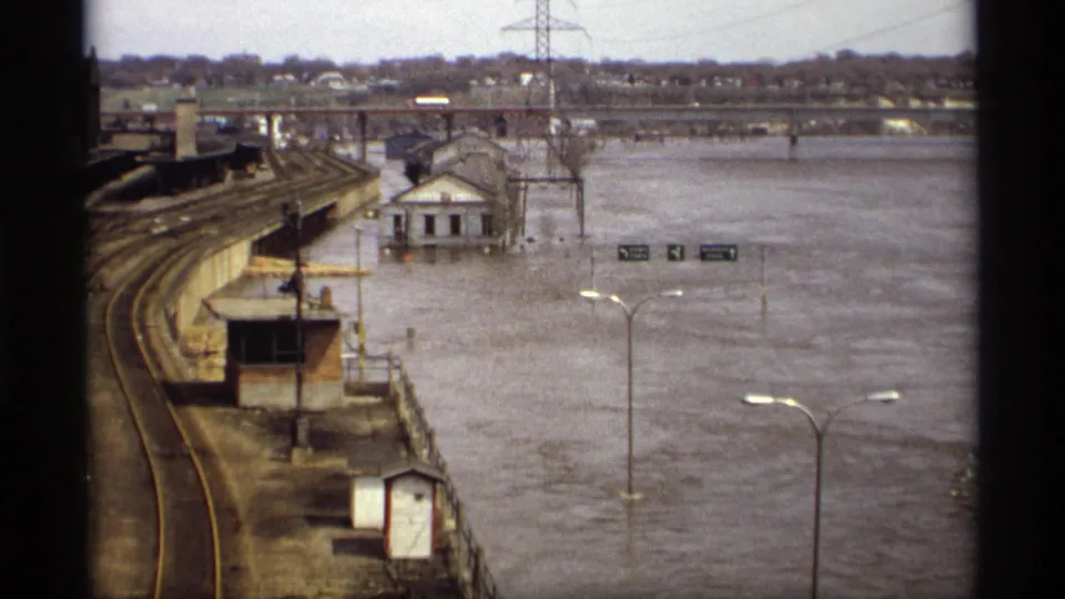 Wide 1969 Saint Paul flood view showing submerged riverfront roadways, rail tracks, streetlights, and a small building near high water.