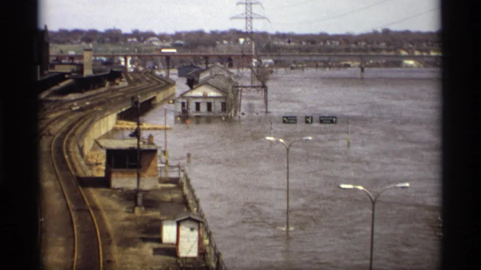 Saint Paul flood scene from an elevated home-movie viewpoint, with rail lines, streetlights, utility structures, and water covering the lower road area.
