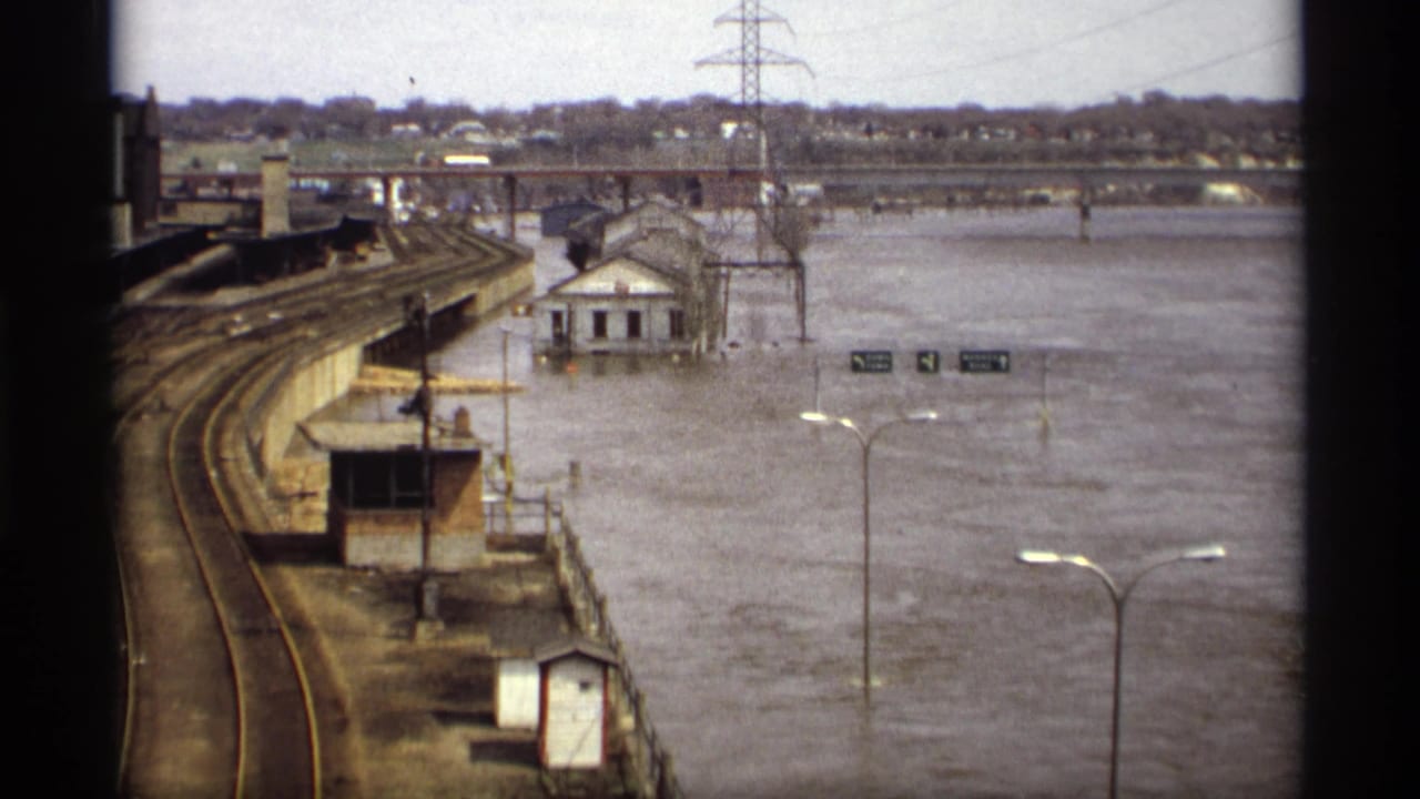 1969 Saint Paul flood scene with curving railway tracks, a small white building in water, streetlights, signs, bridge, and power lines.