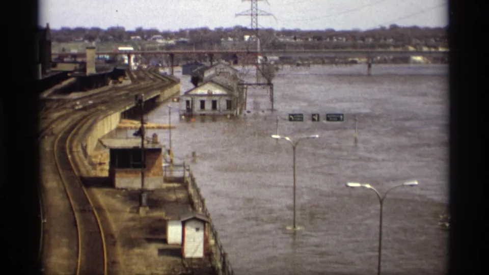 Saint Paul flood footage shows rail tracks beside high water, a small white riverside building, road signs, streetlights, bridge, and power lines.