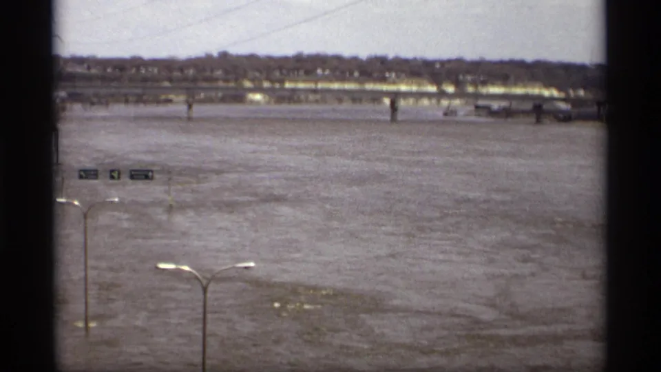 Saint Paul flood scene looking over high water toward a distant bridge and riverside buildings, with traffic signs and streetlights in the foreground.