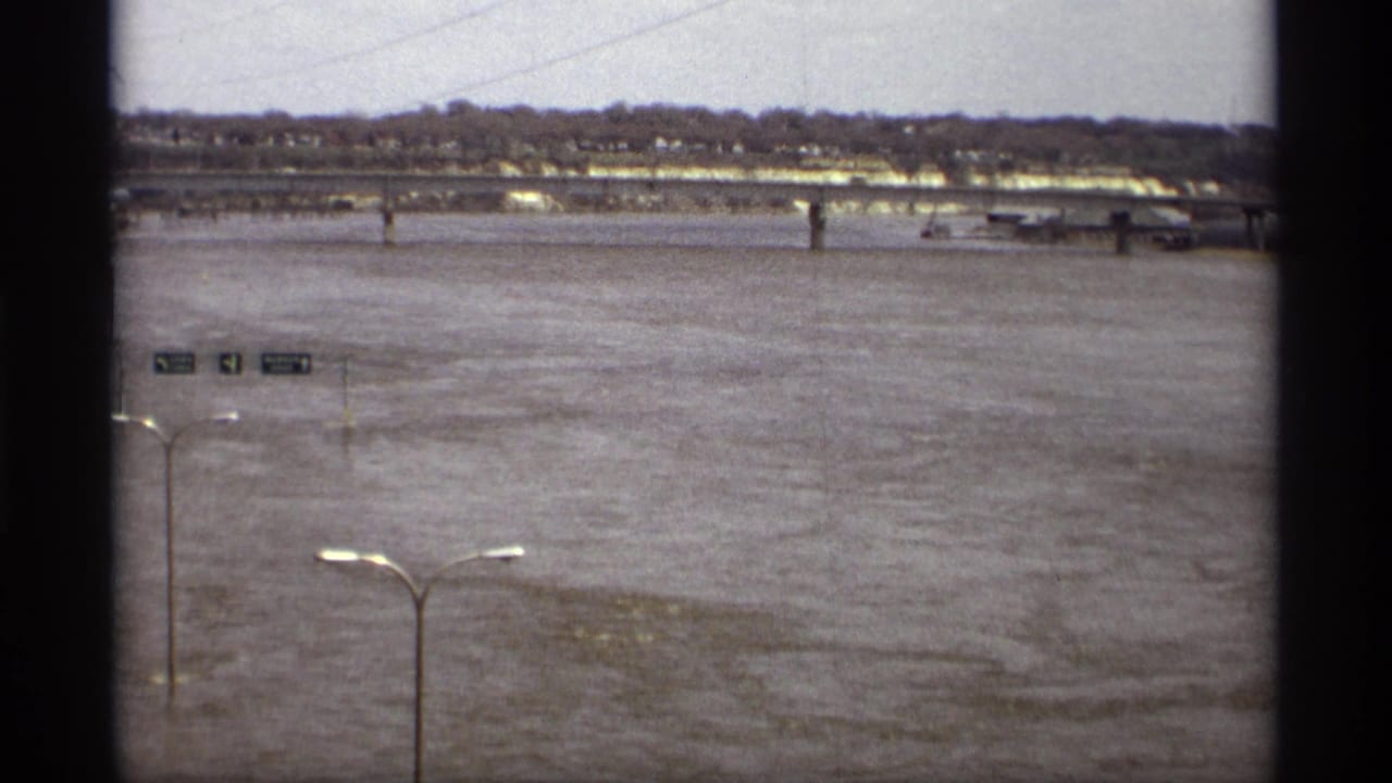 1969 Saint Paul 8mm view of a flooded river, distant bridge, shoreline trees, and partially submerged roadside signs.