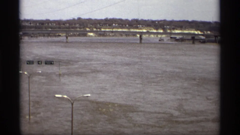 1969 Saint Paul 8mm view of a flooded river, distant bridge, shoreline trees, and partially submerged roadside signs.
