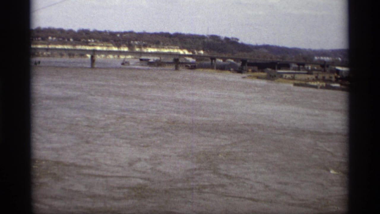 Wide flooded river with bridge, shoreline, and low buildings in the distance.
