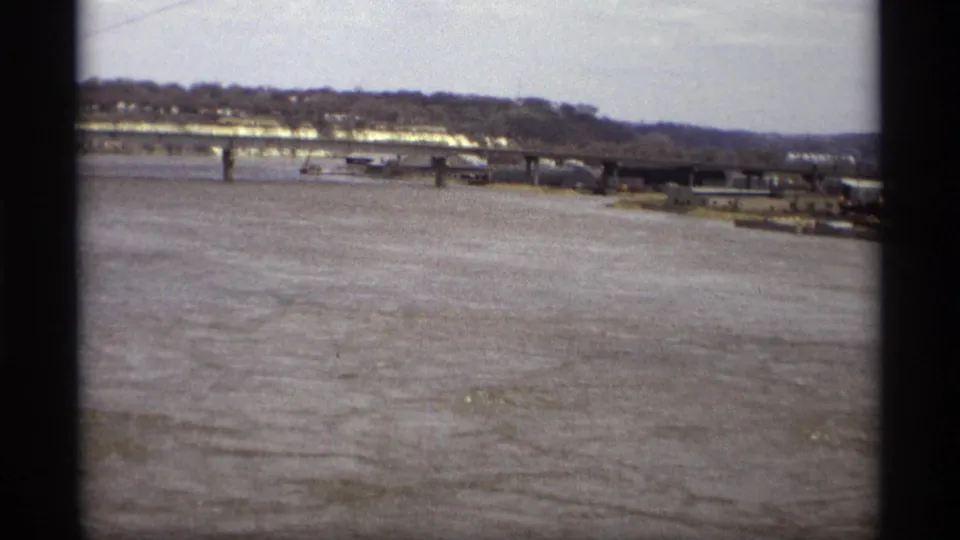Home-movie frame showing Saint Paul floodwater, a distant bridge, shoreline trees, and riverfront structures through a dark vertical edge.