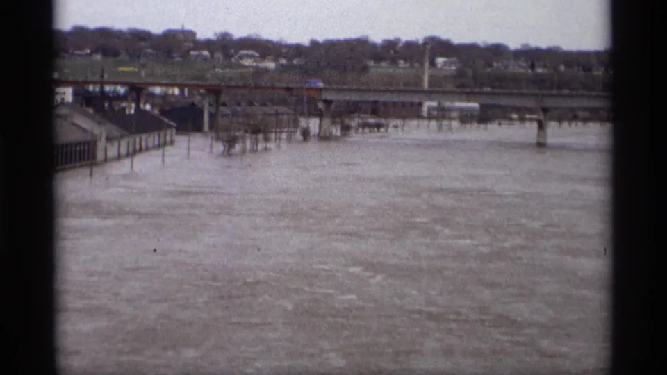 1969 Saint Paul home-movie frame showing a wide flooded riverfront or roadway area beneath a bridge with distant houses under cloudy light.