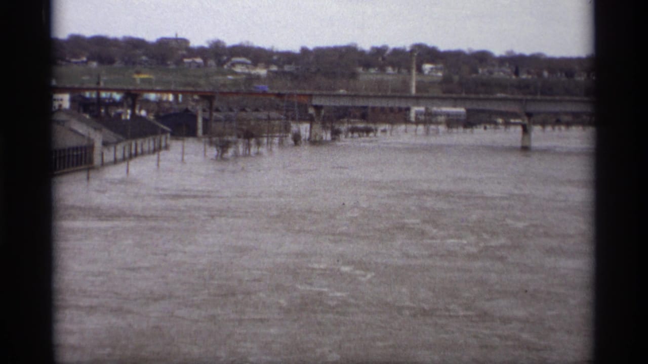 Muddy floodwater flows beneath a bridge with distant houses and leafless trees beyond the river.