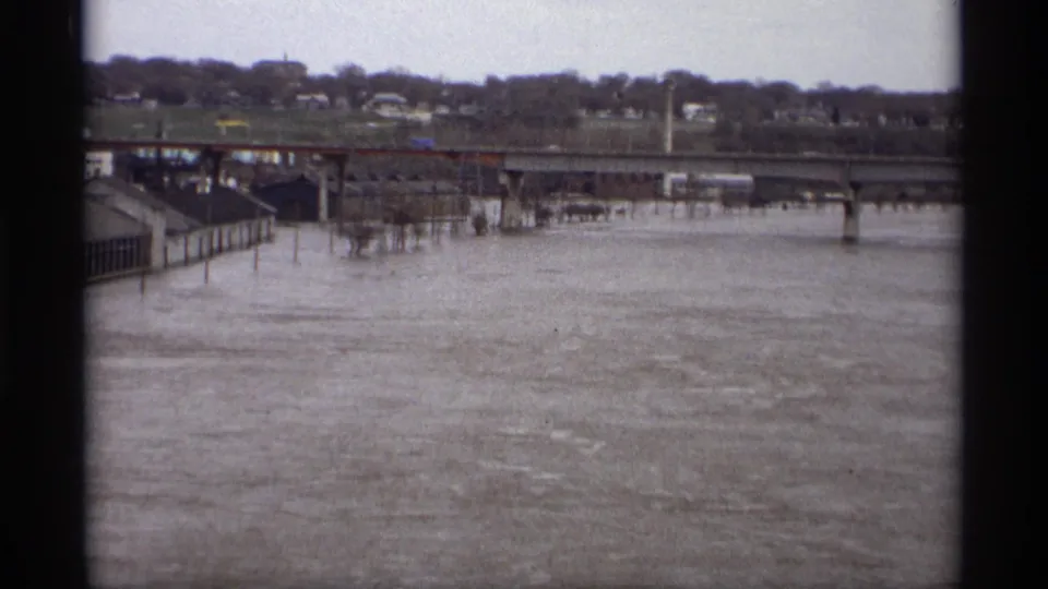 Saint Paul, Minnesota 1969 home-movie frame showing muddy floodwater, a bridge, riverfront structures, and distant houses under cloudy light.