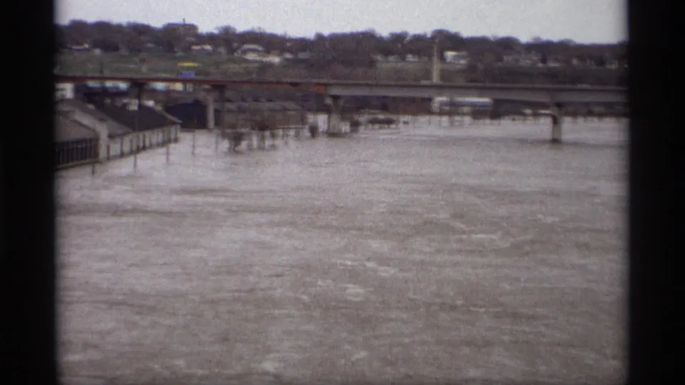 Saint Paul, Minnesota 1969 frame showing high muddy river water below a bridge, with concrete riverfront structures and distant houses.