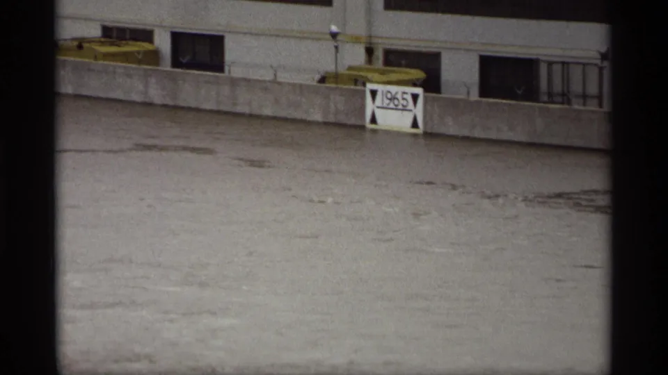 Saint Paul, Minnesota 1969 frame showing muddy floodwater, a concrete wall, a 1965 marker sign, and riverfront building details.