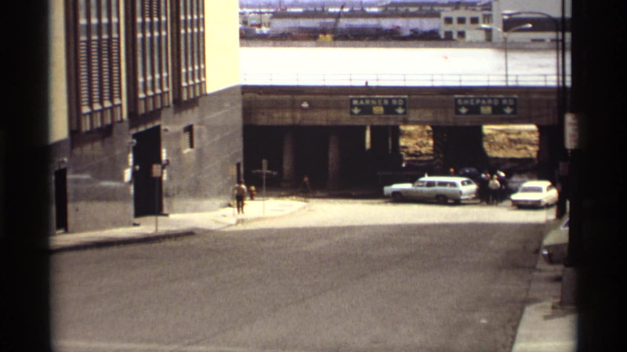 Street leading toward an underpass with direction signs, parked cars, and a distant pedestrian.