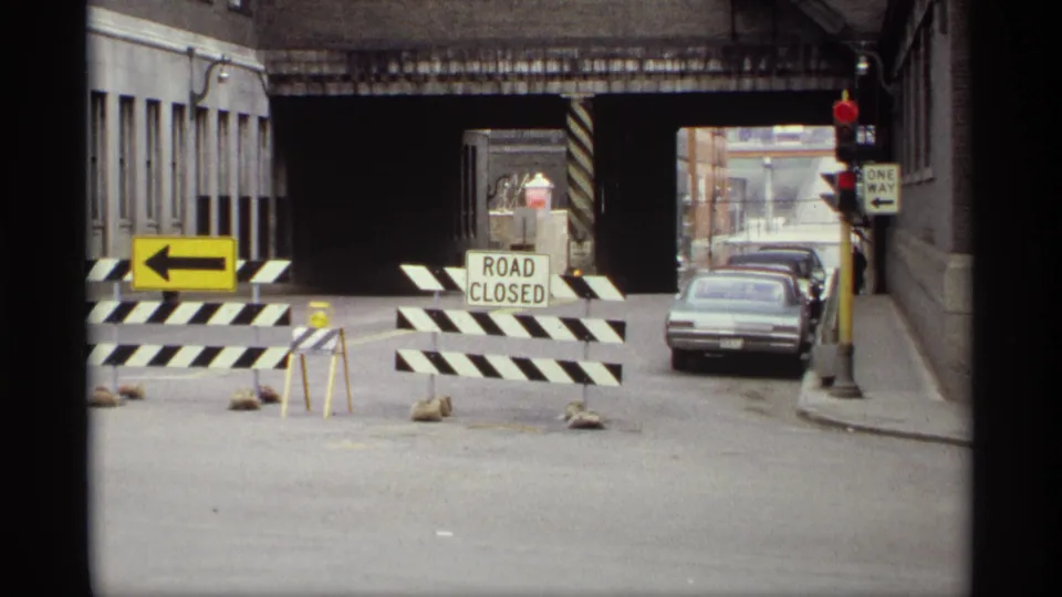 1969 Saint Paul road closure scene with striped barricades, a ROAD CLOSED sign, grey sedan, traffic light, and overpass structure.