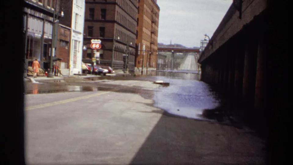 Saint Paul, Minnesota street in 1969 with standing water, parked late-1960s cars, brick buildings, and a Route 66 sign near the corner.