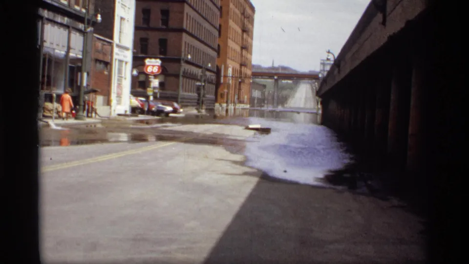 1969 Saint Paul view of a wet street with a large puddle, a visible Route 66 sign, brick buildings, parked cars, and bridge shadow.