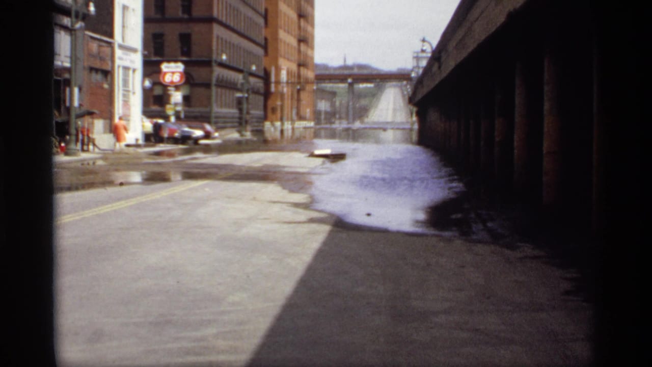 Flooded urban street near brick buildings with a Route 66 gas sign and parked cars.