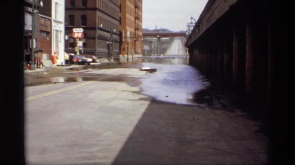 Saint Paul street flooding in 1969, with brick industrial buildings, parked cars, and a Route 66 gas station sign visible beyond the water.