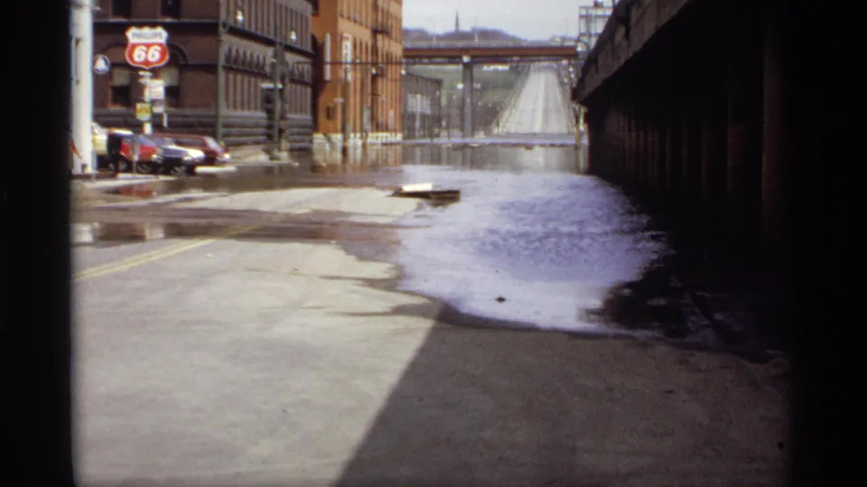 1969 Saint Paul floodwater covers an industrial street near a Route 66 gas sign, with cars at the curb and a bridge in the distance.