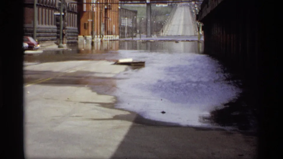 Saint Paul flood scene from 1969 showing water across the street, a floating wooden pallet, brick buildings, and a distant bridge.