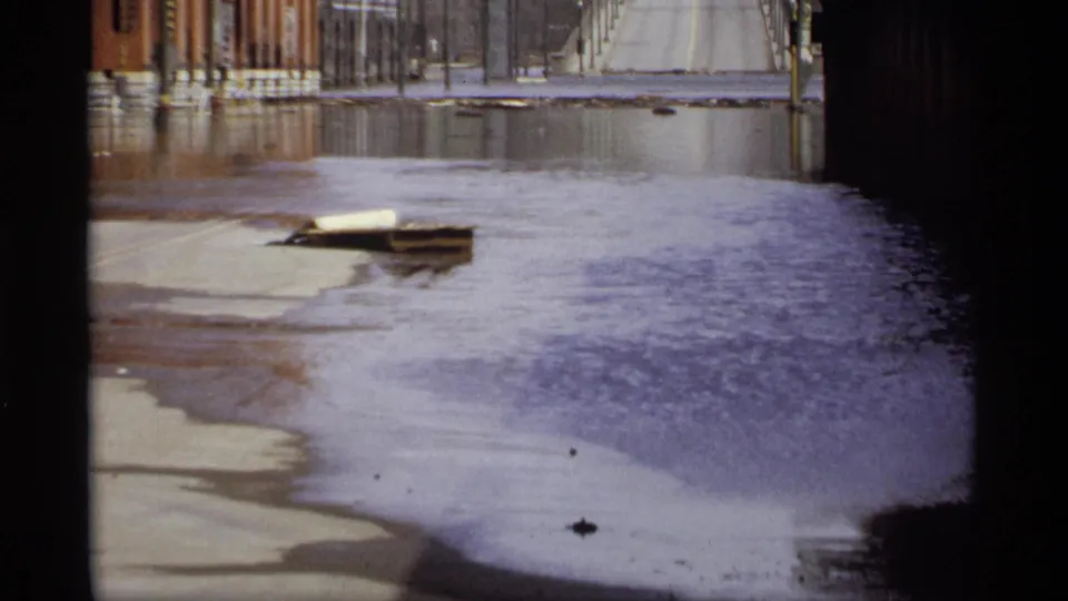 1969 Saint Paul floodwater fills an industrial street around a floating wooden pallet, with brick buildings and a distant bridge visible.