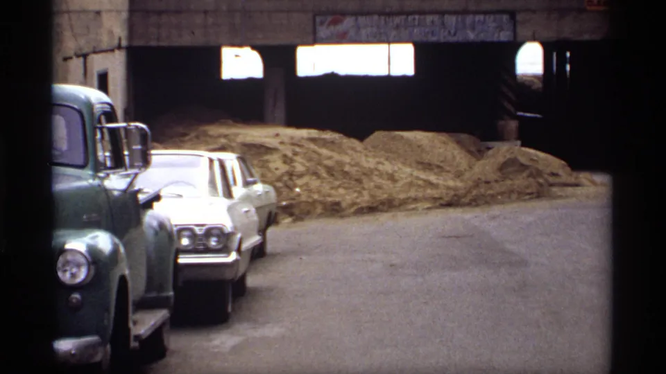 Saint Paul 1969 home-movie frame showing parked classic vehicles beside hay or straw piles beneath a concrete overpass.