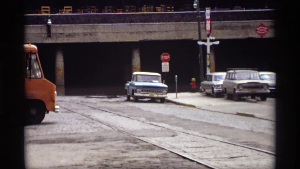 1969 Saint Paul street scene with streetcar tracks, a stop sign, parked older cars, an orange truck, and a concrete underpass.