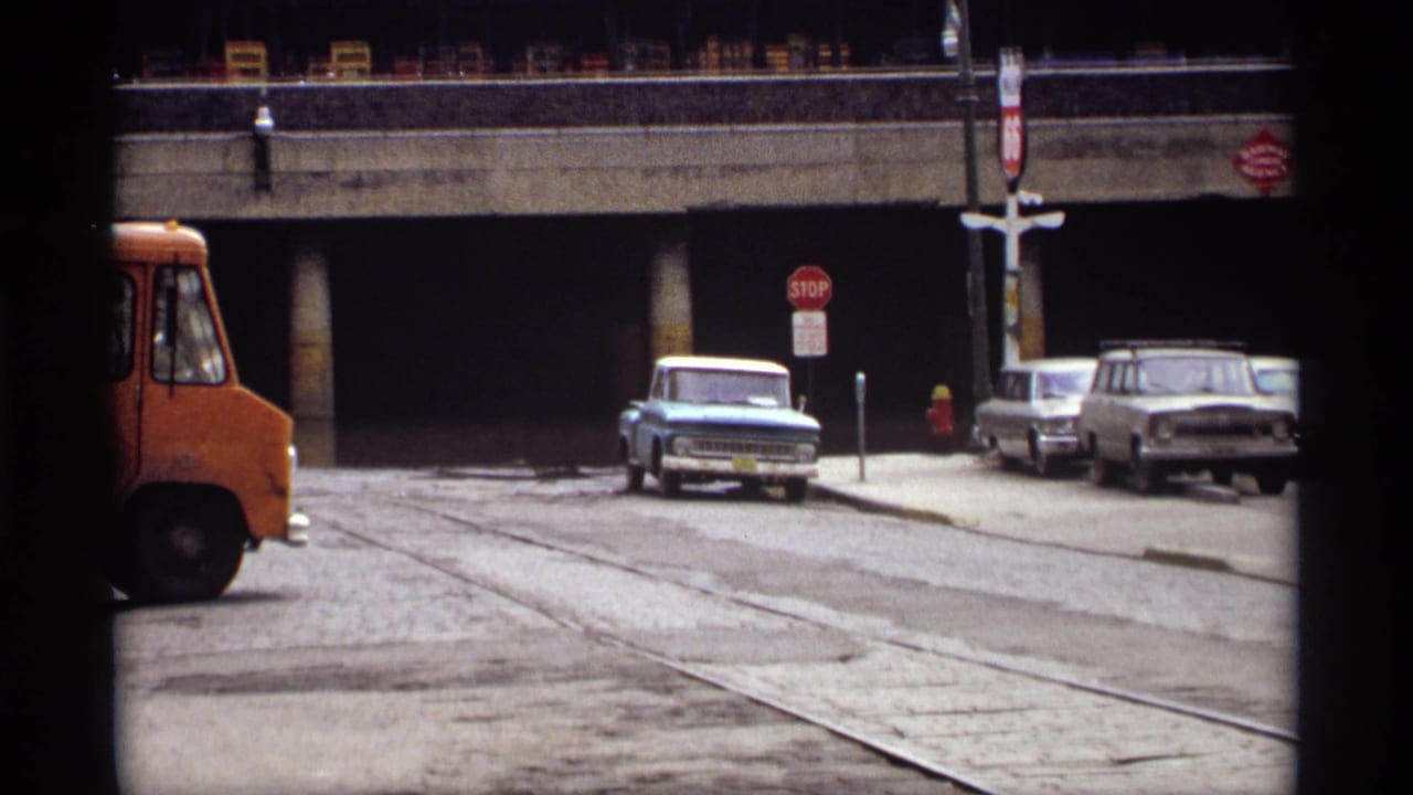 1969 Saint Paul street view beneath a concrete overpass, with parked cars, a stop sign, and the front of an orange vehicle entering from the left.