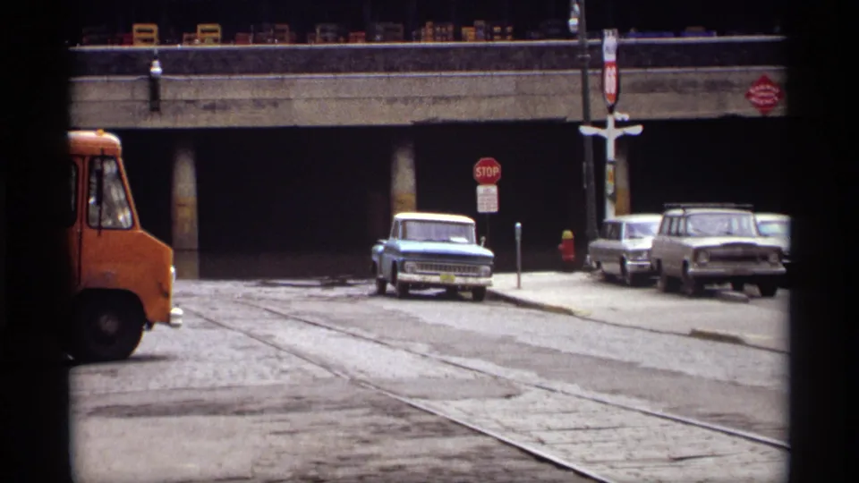 1969 Saint Paul street scene showing a light blue pickup, parked cars, a stop sign, and shadowed space beneath a concrete overpass.