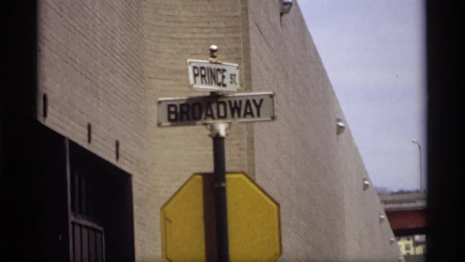 1969 Saint Paul view of Broadway and Prince St street signs mounted above a yellow sign near a brick building wall.