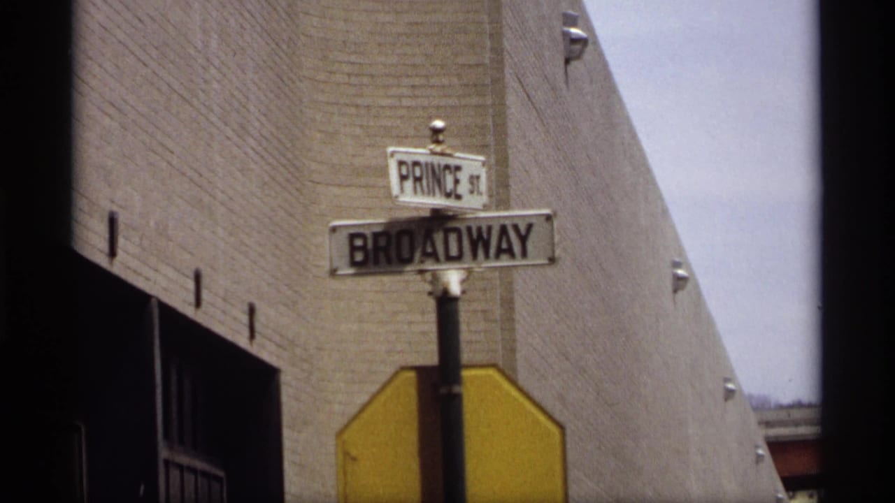 Prince Street and Broadway signs above a yellow stop sign beside a brick wall.