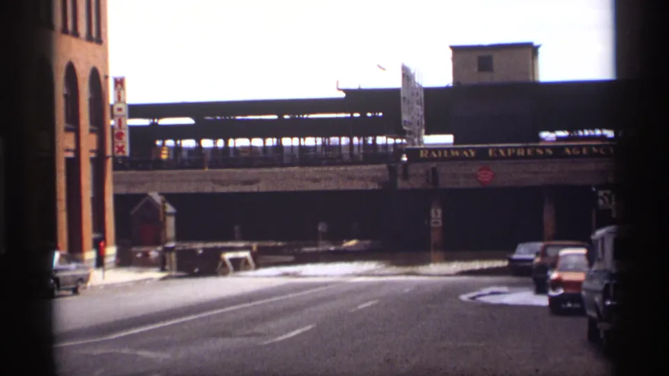 A train passes across the elevated railway bridge above a Saint Paul street lined with late-1960s cars and commercial signage.