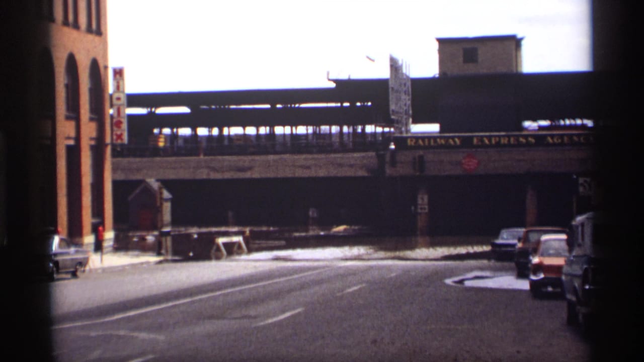 Saint Paul 1969 flood scene showing water across a street, classic cars, brick buildings, and a Railway Express Agency sign under elevated rail tracks.