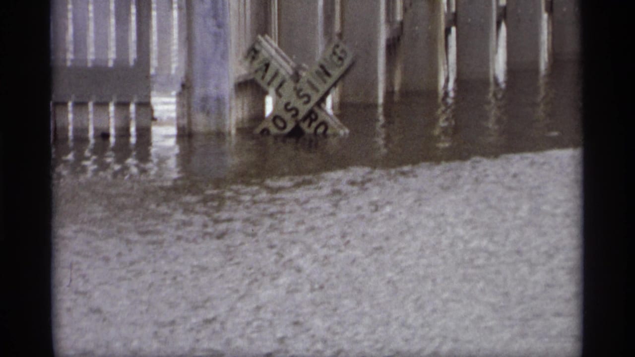 1969 Saint Paul floodwater rises around a railroad crossing sign and nearby pilings, with rippled water filling the foreground.