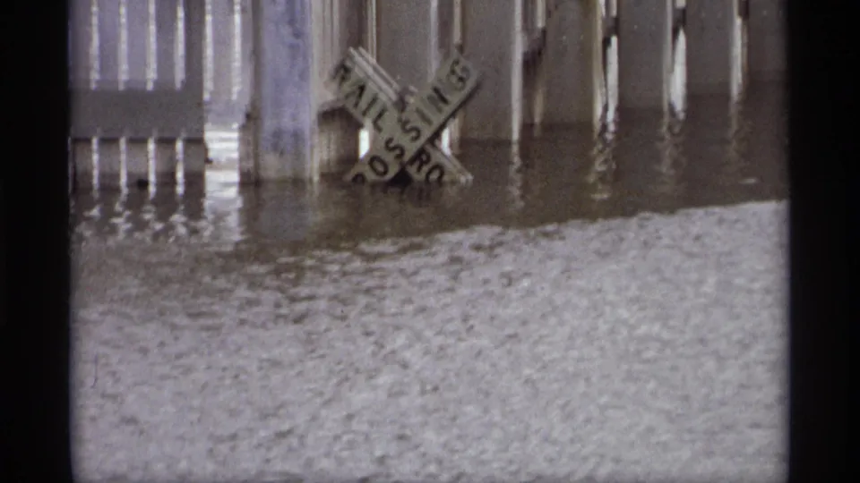1969 Saint Paul floodwater rises around a railroad crossing sign and nearby pilings, with rippled water filling the foreground.