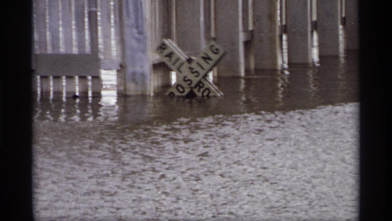 A railroad crossing sign leans in floodwater next to pilings and a partially submerged barrier.