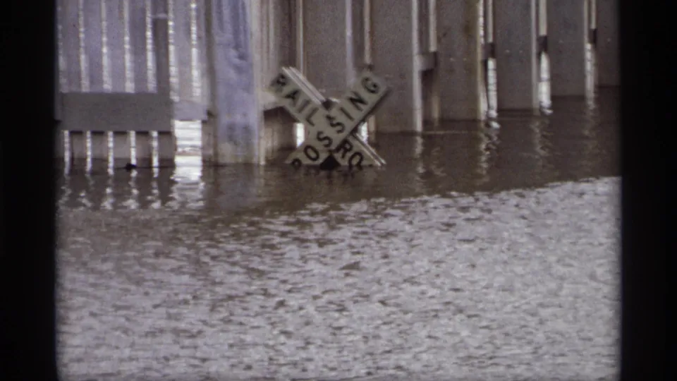 In the Saint Paul flood scene, the crossbuck sign appears tilted as high water surrounds the crossing area and pilings.