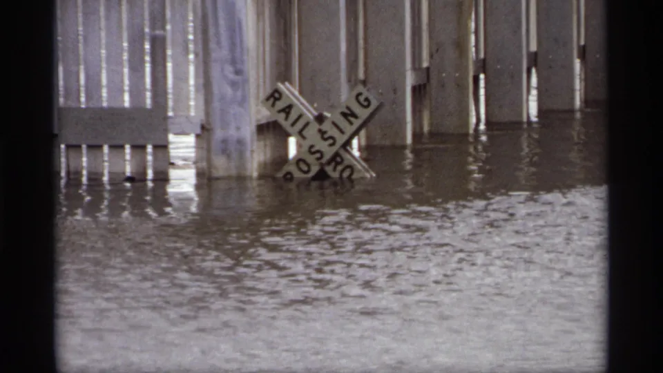 A close view shows the Saint Paul railroad crossing sign partly submerged by floodwater, with pilings and reflections behind it.