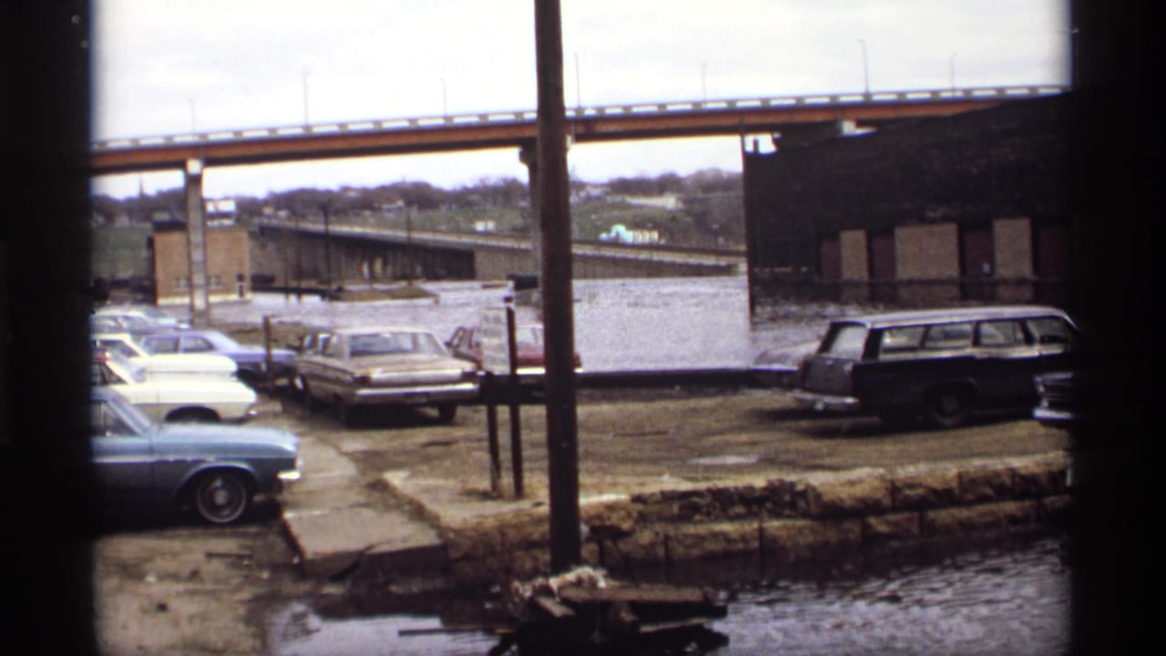 Flooded parking area with classic cars, bridge, brick building, and dark window frame.